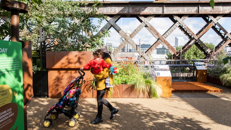 Woman in a yellow jacket holding her daughter who is wearing a red jacket and pushing a buggy along a path surrounded by large metal planters filled with plants and trees
